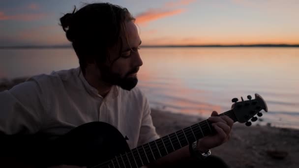 Gedachte bebaarde hipster man die een akoestische gitaar speelt bij het meer tegen een zonsondergang hemel - slow-motion portret shot — Stockvideo