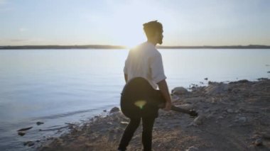 Gitarlı hippi, göl kıyısında günbatımının tadını çıkarır. Taşlı sahilde güneşe karşı yürür.