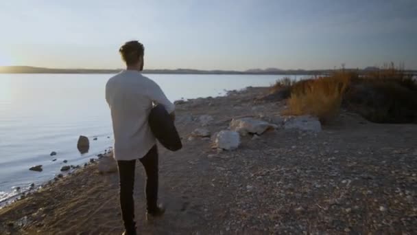 Baksidan av en hipster man promenader längs stranden mot solen håller en gitarr, Torrevieja Rosa sjön i Alicante, Spanien — Stockvideo
