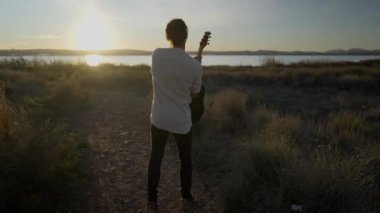 Genç bir gitaristin, elinde akustik bir gitarla gün batımına doğru kollarını kaldırması. Yavaş çekim.