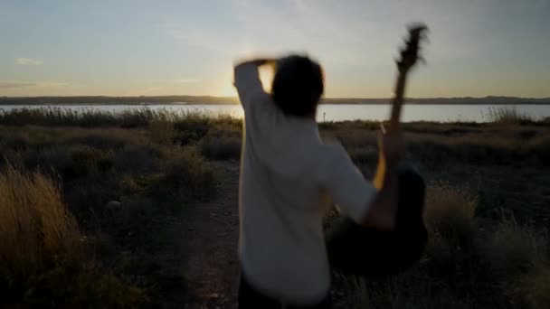 Vue arrière de Man sautant avec une guitare regardant le coucher du soleil sur le lac — Video