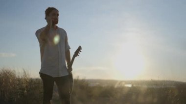 Genç sakallı gitaristin portresi, günlük beyaz gömlekli cep telefonuyla konuşan, elinde gitar tutan açık havada gölün üzerinde gün doğumuna bakan bir adam.