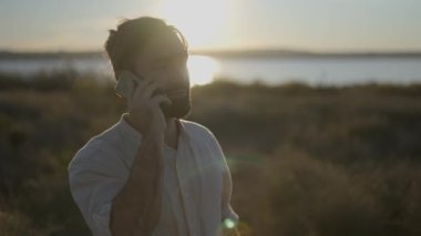 Cep telefonuyla konuşan genç adam, günbatımı ışığıyla arka planda.