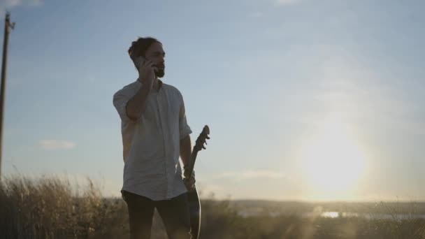 Homem de pé com uma guitarra na mão e falando ao telefone ao pôr do sol — Vídeo de Stock