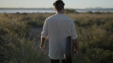 Hipster bir adamın elinde gitarla gün batımında çimenli bir tarlada yürüyor. 