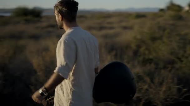 Ισπανός hipster άνθρωπος με μια κιθάρα που περπατά μέσα από ένα χορτώδες πεδίο προς το ηλιοβασίλεμα - μετά από gimbal shot — Αρχείο Βίντεο