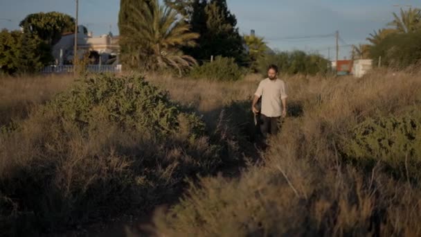 Spanska skäggiga killen går längs gräsplan väg med akustisk gitarr i handen — Stockvideo