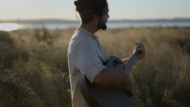 Man Guitarist De pie en el campo de hierba en España y tocando acordes en la guitarra acústica negra contra la hermosa luz del sol - vista de perfil en cámara lenta — Vídeo de stock
