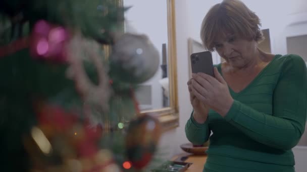 Mulher espanhola velha em casa tirando foto da decoração da árvore de Natal com câmera Smartphone. tiro médio — Vídeo de Stock