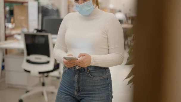 Bizneswoman w masce medycznej twarzy przy użyciu telefonu SMS z obu rąk w środowisku biurowym — Wideo stockowe