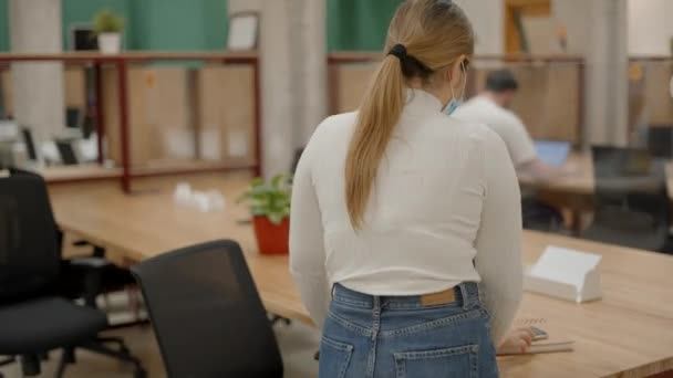 En ung iværksætter med en beskyttende ansigtsmaske sætter sig på sin arbejdsplads i et kollaborativt kontor under covid-19 pandemien. Young Business Woman sidder ved skrivebordet foran laptop – Stock-video