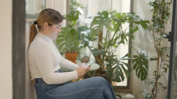 Kaukasiske smilede munter kvinde sidder på en stol med en smartphone i hænderne og hvile. Kvindelig aflytning, rulning og browsing online. Lady ser noget på telefonen. – Stock-video
