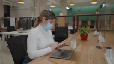 Koruyucu yüz maskeli bir kadın, birlikte çalıştığı ofisteki dizüstü bilgisayarla online ödeme için kredi kartı kullanıyor. 