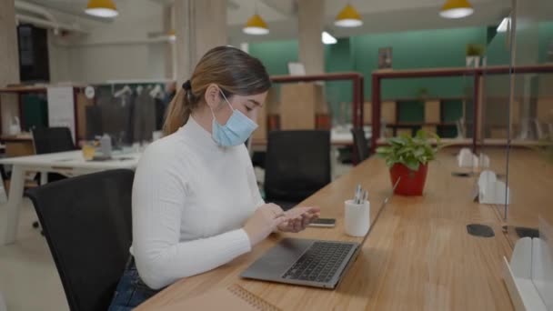 Mujer con mascarilla protectora usando tarjeta de crédito para pago en línea con computadora portátil en la oficina de coworking — Vídeos de Stock