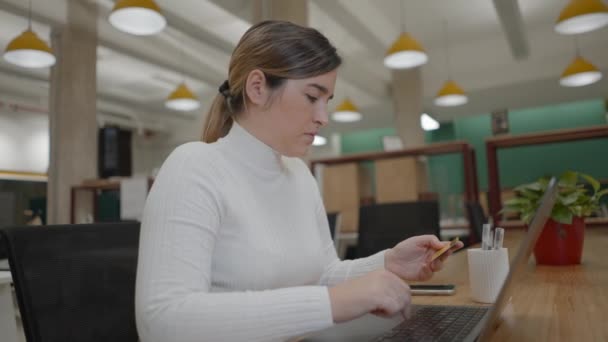 Mujer joven seria y exitosa sentada en el espacio de coworking, sosteniendo una tarjeta de crédito en su mano escribiendo números o ingresando código de pin, mirando atentamente a la pantalla del portátil, haciendo pedidos en línea — Vídeos de Stock