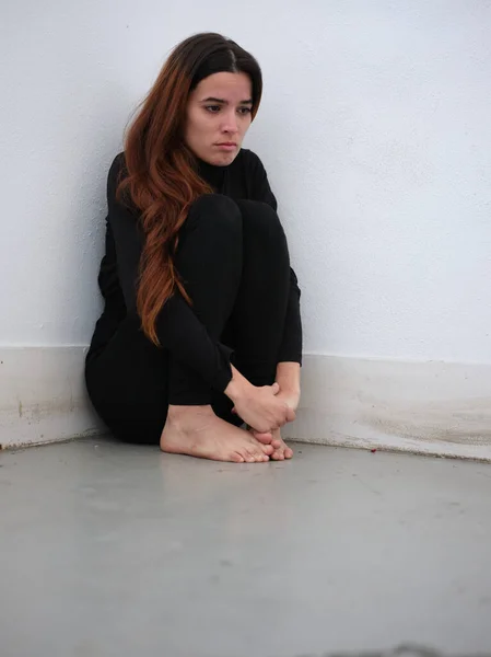 Baixo ângulo vista de uma jovem mulher no canto de uma sala vestida de preto descalço olhando triste — Fotografia de Stock