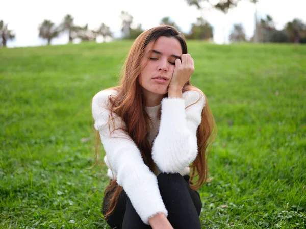 Uma jovem mulher no parque sentada na grama com uma mão no rosto adormecendo. — Fotografia de Stock