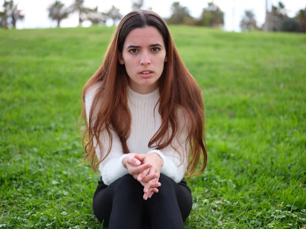 Uma jovem irritada no parque sentada na grama com as mãos juntas olhando para a câmera. — Fotografia de Stock