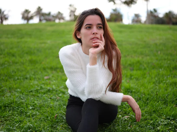 Uma jovem mulher pensativa sentada na grama no parque apoiando o rosto com a mão e olhando para longe — Fotografia de Stock