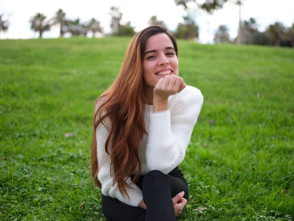 Uma jovem apaixonada sentada na grama do parque sorrindo para a câmera com a mão no queixo. — Fotografia de Stock