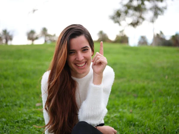 Jag har en idé. En ung kvinna i parken tittar på kameran leende och höjer sitt pekfinger. — Stockfoto