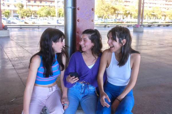 Tři přátelé se smějí při používání mobilu sedí na městském prostoru — Stock fotografie