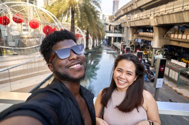 Afro adam ve Asyalı kadın dışarıda selfie çekerken kameraya gülümsüyor.
