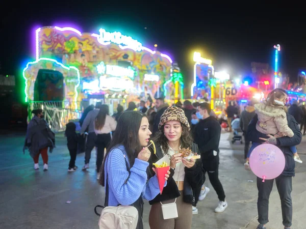 Two women eating fast food in a crowded and colorful night fair — Stock Photo, Image