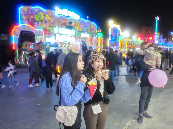 Women eating waffle and fries standing in the middle of a crowded night fair — Stock Photo, Image