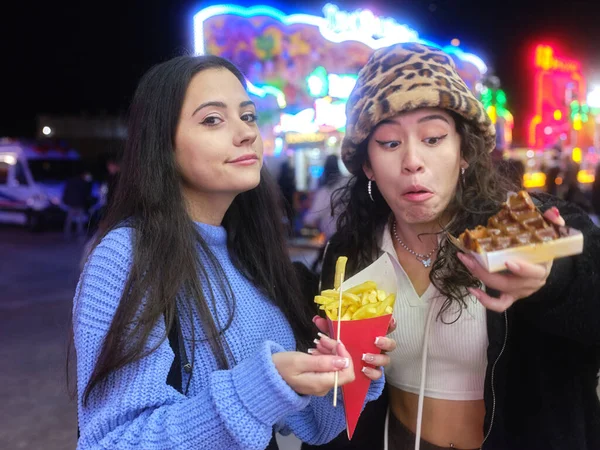 Woman hesitantly looking at a waffle next to a friend with fries in a night fair — Stock Photo, Image