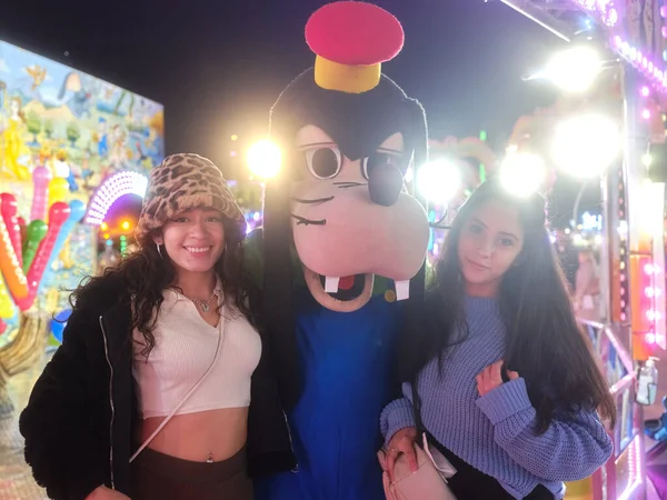 Дві жінки-латиноамериканки стоять поруч з актором, одягненим як собака на нічному ярмарку — стокове фото