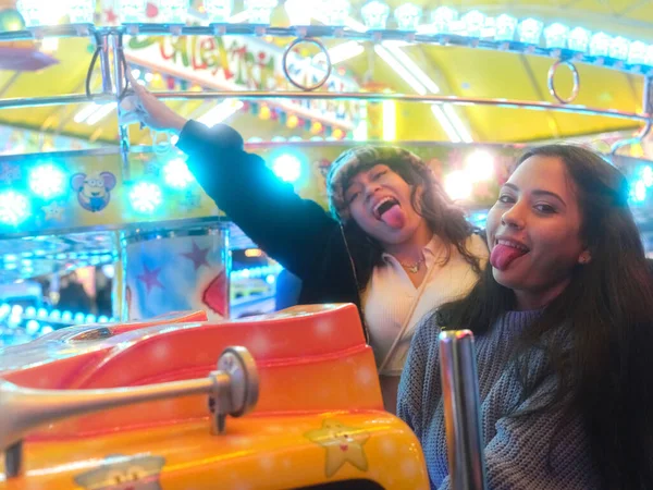 Two woman grimacing with the tongue out inside a carousel car in a night fair — Stock Photo, Image