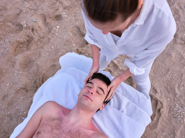 Молодой человек, лежащий на массажном столе и получающий массаж головы на пляже в Валенсии от молодого специалиста по хиромассажу. — стоковое фото
