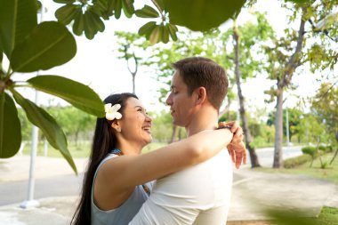 Saçında beyaz tropikal çiçekli bir kadın parkta bir adamı kucaklıyor.