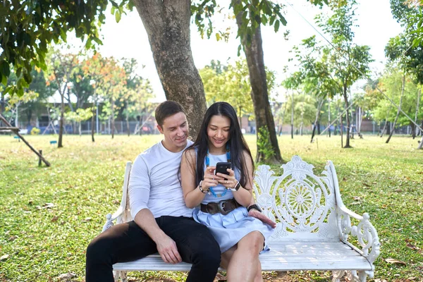 Multiethnic couple sitting together on a bench while using the mobile outdoors — Foto Stock