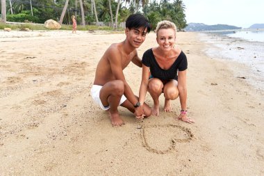 Kumsalda çömelip kuma kalp çizen çift.