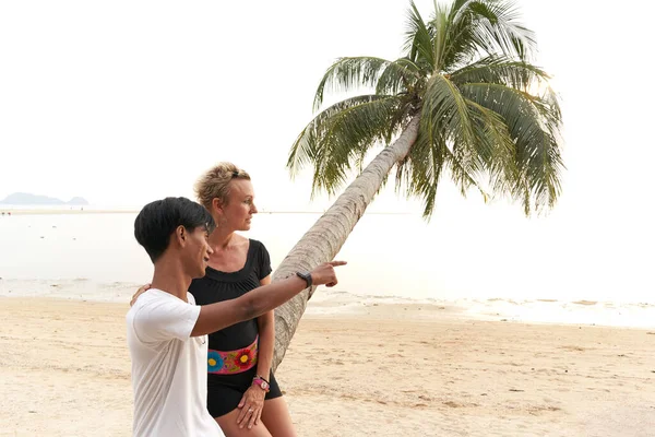 Young asian man pointing ahead next to a woman sitting on a palm tree — ストック写真