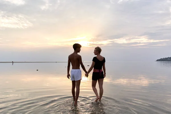 Multiethnic couple holding hands and walking into the sea during sunset — ストック写真