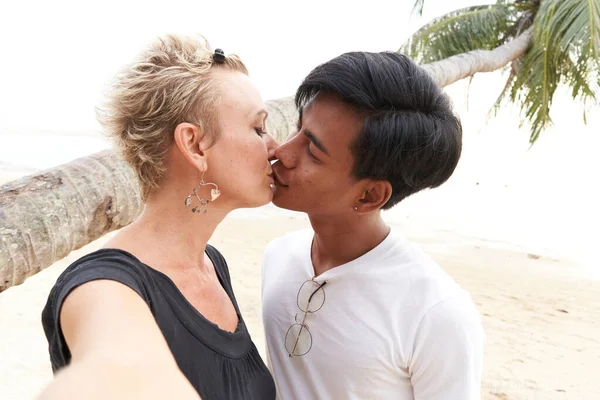 Multiracial lovers kissing and taking a selfie on the beach — ストック写真