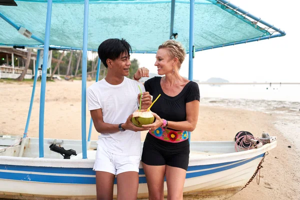 Multicultureel koppel zittend op een aangemeerde boot sap drinkend van kokosnoot — Stockfoto