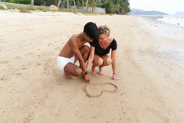 Multicultureel echtpaar op een strand tekenen een hart vorm samen op het zand — Stockfoto
