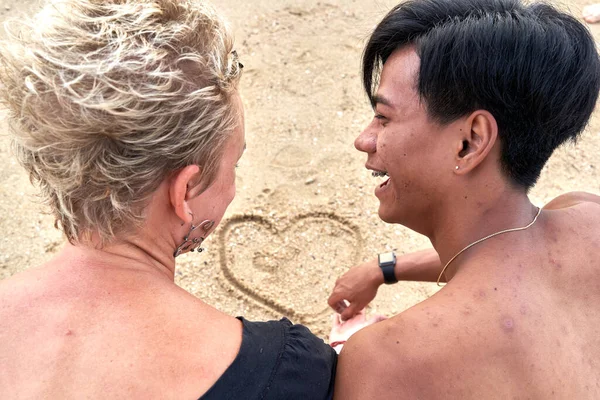 Back of a multiethnic couple drawing a heart shape on the sand of a beach — ストック写真