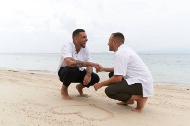 Tropik bir plajda kumlara çizilmiş bir kalbin yanında el ele tutuşan eşcinsel çift.