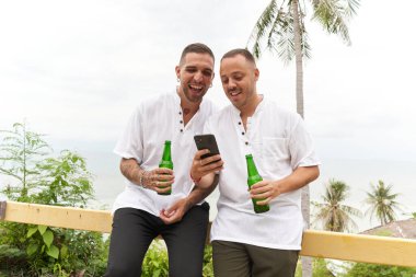 Eşcinsel çift deniz manzaralı terasta bira içerken cep telefonunu kullanıyor.
