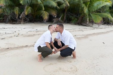 Tropik bir plajda çömelmiş, kumlara kalp çizen eşcinsel bir çift.