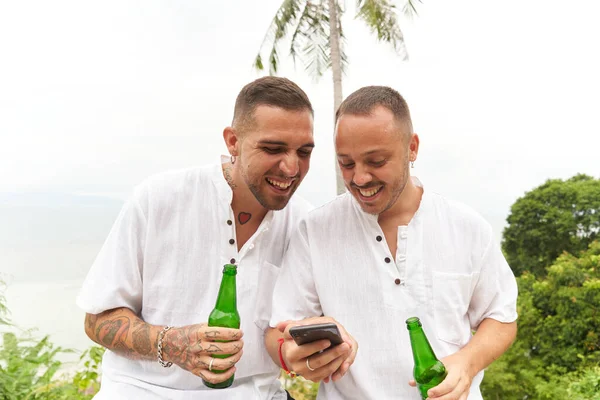 Gay coppia in bianco utilizzando un mobile mentre tenendo birra accanto a un tropicale spiaggia — Foto Stock