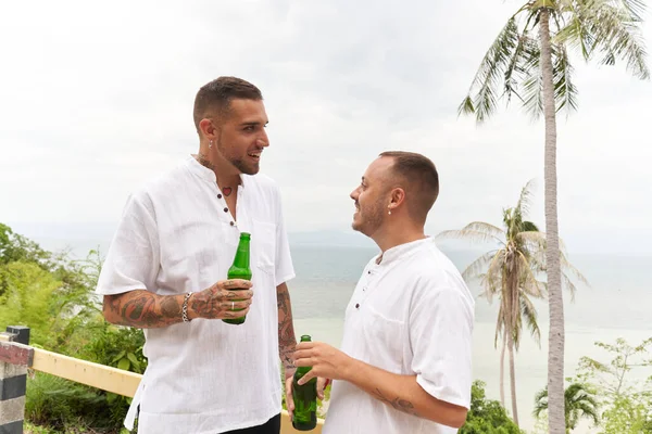 Dois gay homens vestir branco beber cerveja ao lado de um praia — Fotografia de Stock