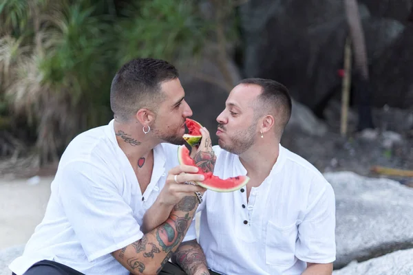 Gay couple joking while feeding each other with watermelon outdoors