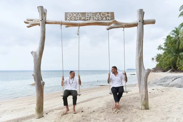 Casal homossexual balançando em um balanço em uma praia tropical — Fotografia de Stock