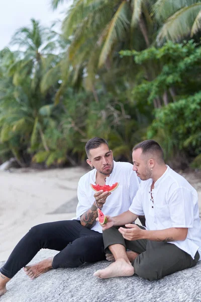 Foto verticale di una coppia gay che mangia anguria su una spiaggia — Foto Stock